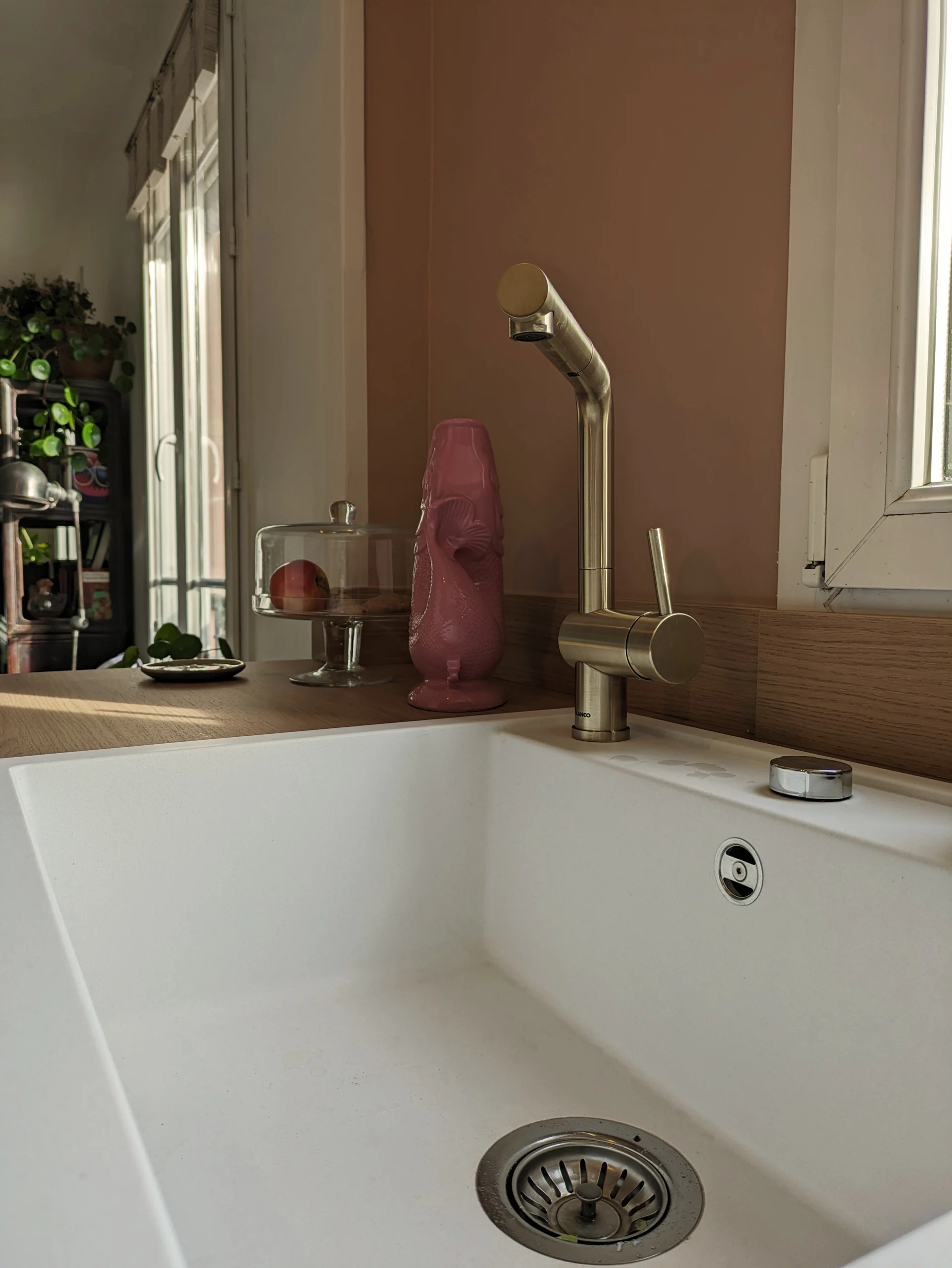 Rénovation de la cuisine - Focus sur l'évier en granit blanc avec un mitigeur doré - arrière plan mur rose Masquerade de Little Greene Focus sur l'évier en granit blanc avec un mitigeur doré - arrière plan mur rose Masquerade de Little Greene et le salon blanc avec des plantes en colonne