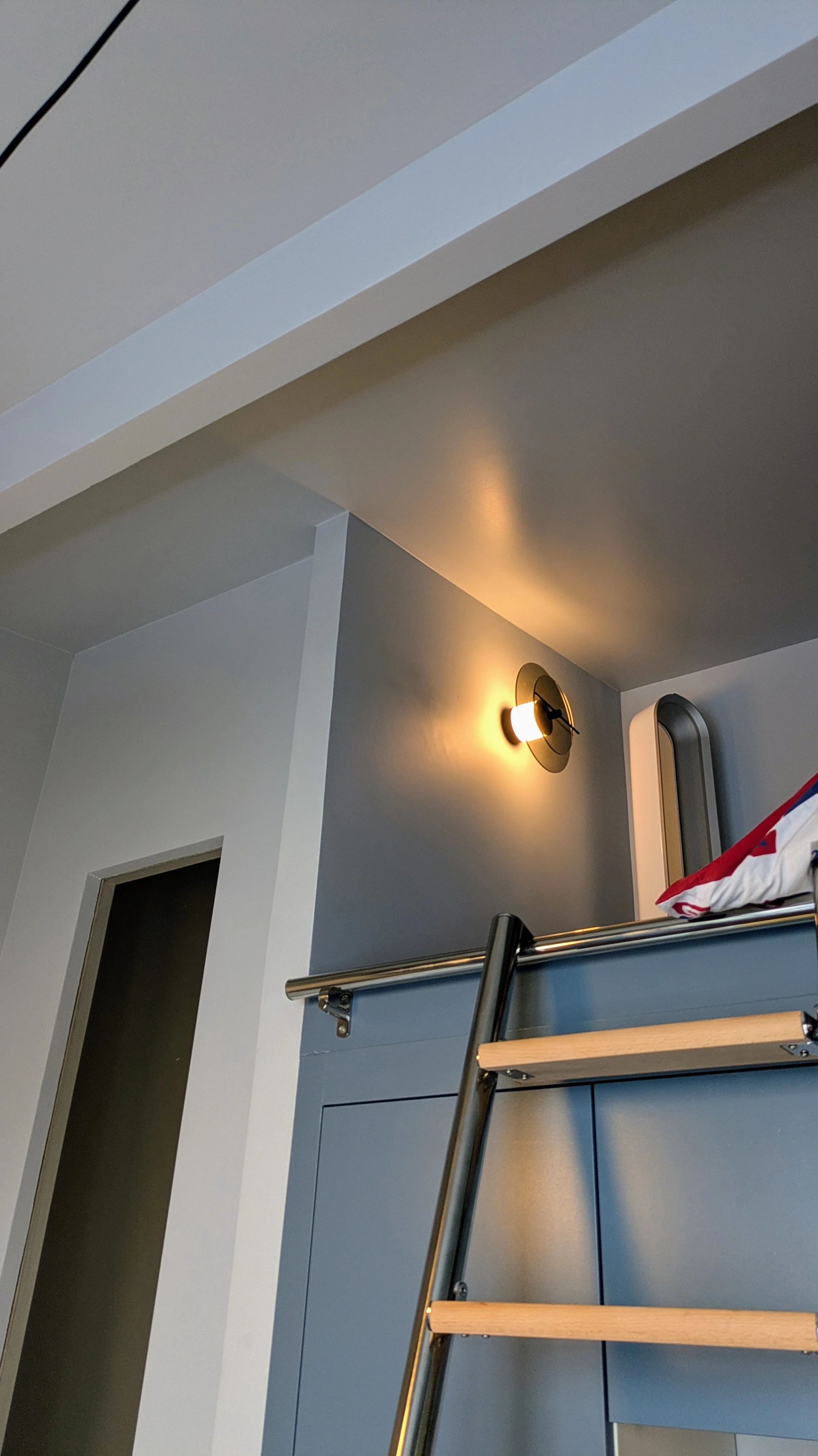 Zoom sur l’applique murale DCW dans cette chambre sous les combles, avec une peinture bleu intense, des poutres laissées apparentes et une ambiance feutrée soigneusement travaillée. Applique murale design DCW dans une chambre rénovée sous toiture, mur bleu profond et poutres apparentes