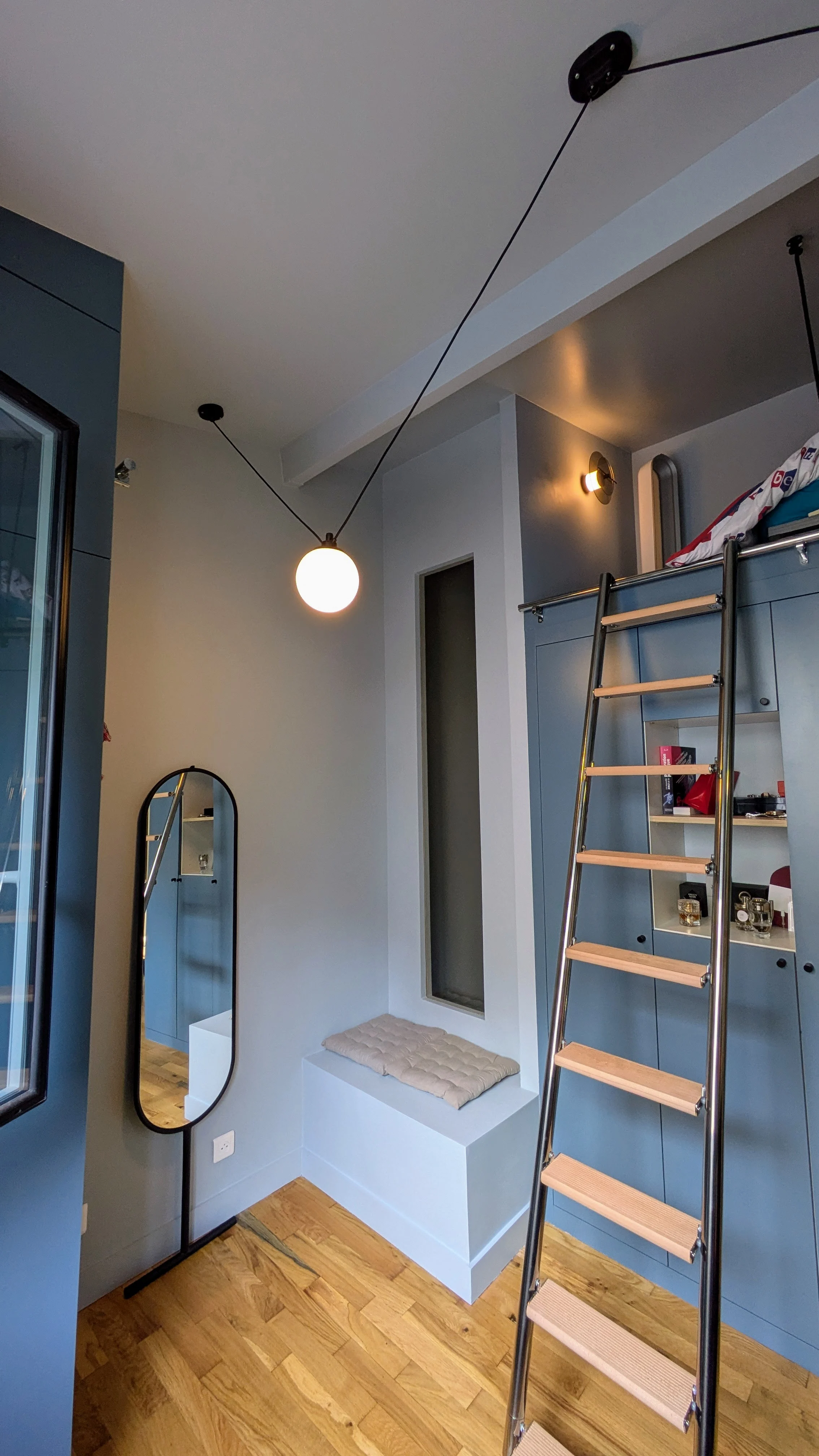 Un coin lecture cocooning niché sous les combles, mis en valeur par une lumière douce et un miroir mural au design contemporain. Espace détente sous les combles cosy, miroir décoratif et éclairage DCW