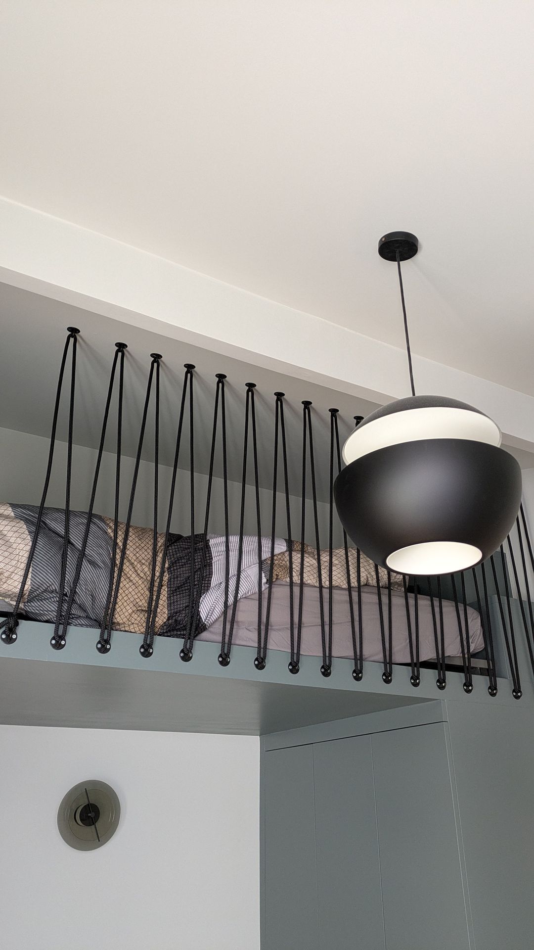 Photo d'un lit mezzanine contemporain intégré, doté d'une balustrade unique composée de multiples cordes noires verticales, offrant à la fois sécurité et un élément de design distinctif. Au-dessus, une suspension moderne bicolore noire et blanche illumine l'espace. Idéal pour optimiser l'espace dans les petites surfaces ou créer une chambre d'enfant stylée. Lit mezzanine moderne avec balustrade décorative à cordes noires et suspension design