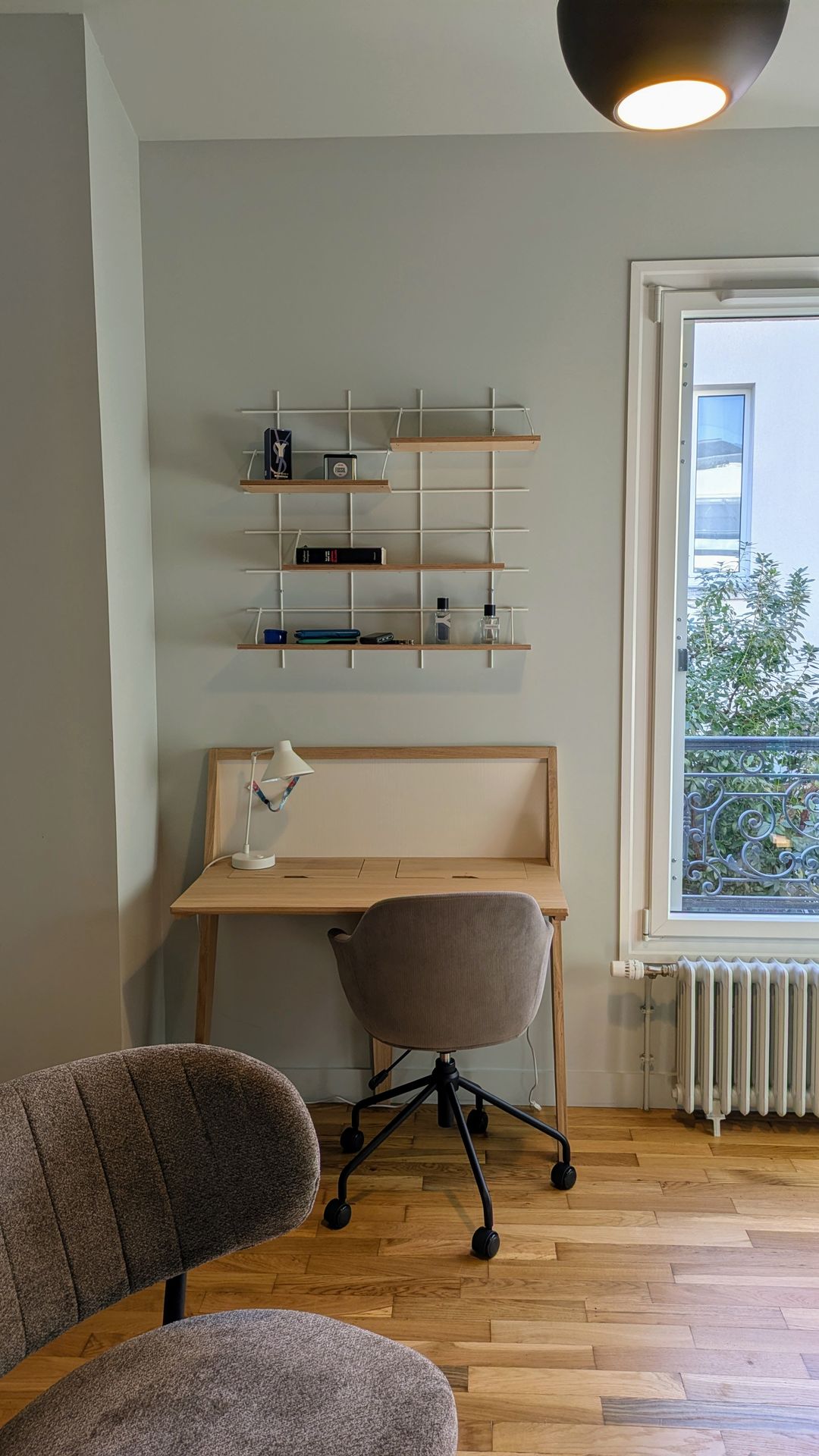 Un espace bureau bien organisé et lumineux dans une pièce, comprenant un bureau en bois clair au design épuré, accompagné d'une chaise de bureau contemporaine sur roulettes. Au-dessus du bureau, une étagère murale grillagée avec des tablettes en bois offre un rangement minimaliste pour des objets et livres. À droite, une fenêtre donne sur l'extérieur, apportant de la lumière naturelle, et un radiateur classique est visible en dessous. Coin bureau moderne en bois clair avec étagère murale grillagée et chaise de bureau à roulettes